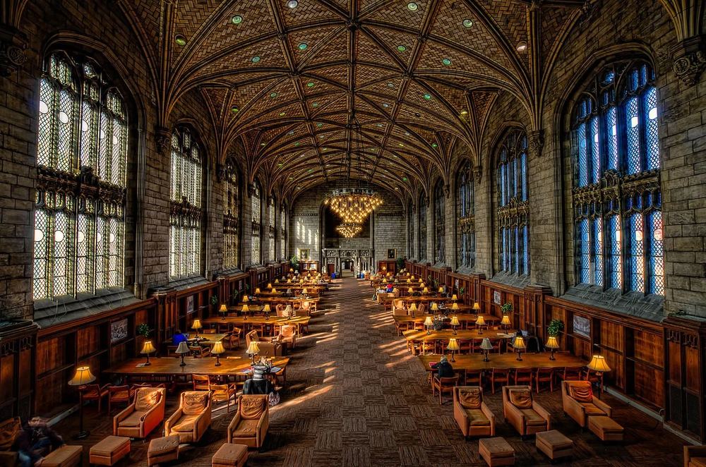 A grand university reading room lined with books and reading lamps